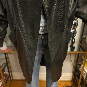 May include: Black leather coat with a textured, patterned panel on the front. The coat has a long length and a classic design. The coat is worn over a denim jacket and blue jeans.