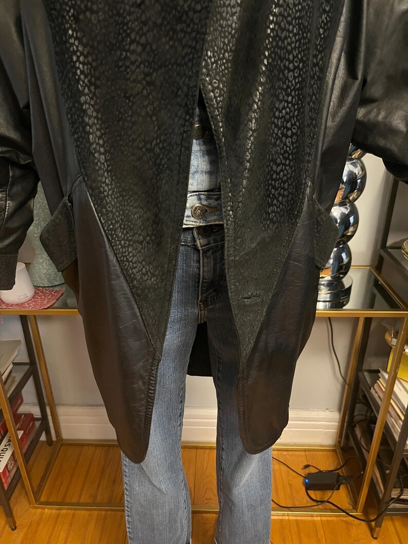 May include: Black leather coat with a textured, patterned panel on the front. The coat has a long length and a classic design. The coat is worn over a denim jacket and blue jeans.