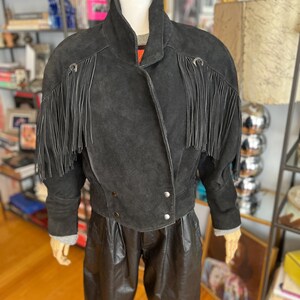 May include: A black suede jacket with fringe detailing on the sleeves and shoulders. The jacket has a high collar and silver button closures. The mannequin is also wearing black leather pants. The jacket is a vintage style.