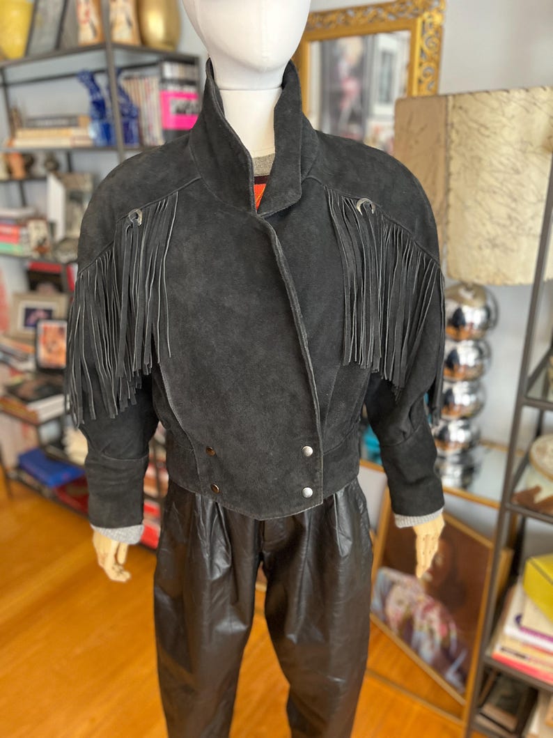 May include: A black suede jacket with fringe detailing on the sleeves and shoulders. The jacket has a high collar and silver button closures. The mannequin is also wearing black leather pants. The jacket is a vintage style.