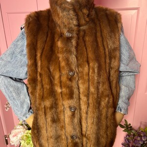 May include: A brown fur vest with a high collar and three buttons down the front. The vest is displayed on a mannequin. The vest is a medium brown color and appears to be made of a soft, plush material.