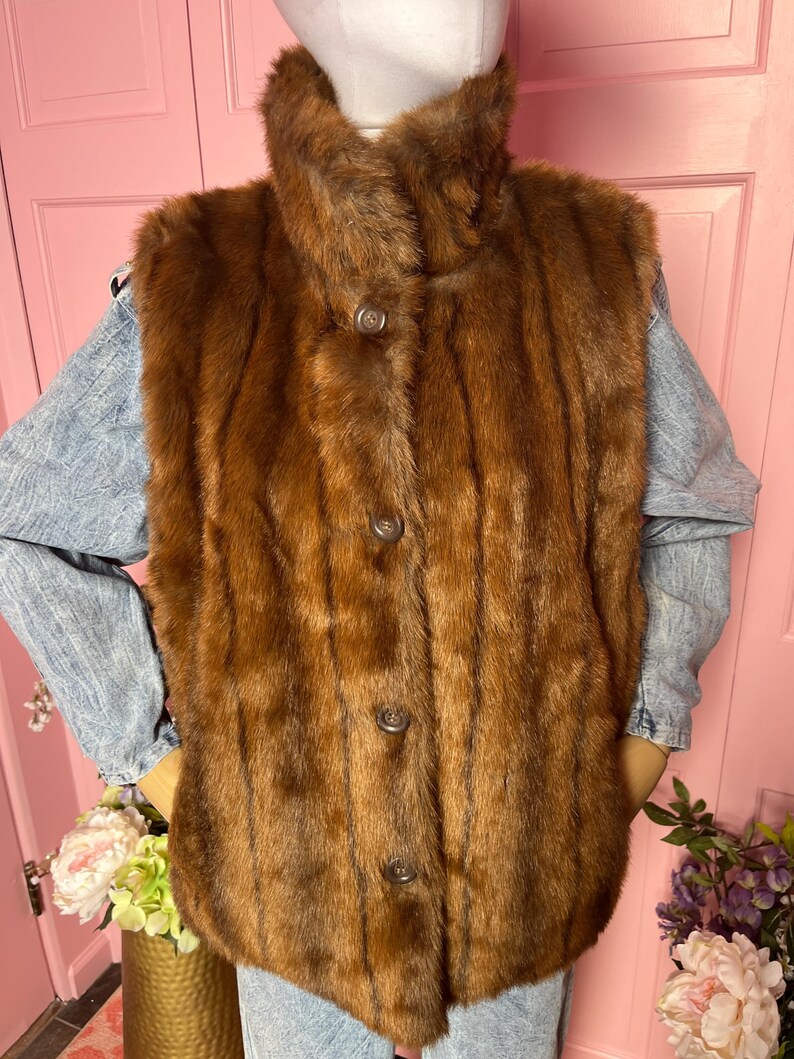 May include: A brown fur vest with a high collar and three buttons down the front. The vest is displayed on a mannequin. The vest is a medium brown color and appears to be made of a soft, plush material.
