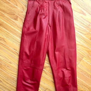 May include: Red leather pants with a high waist and a belt. The pants have pleats at the front and pockets. The leather has a slight sheen. The belt has a buckle closure. The pants are a bold, solid color.