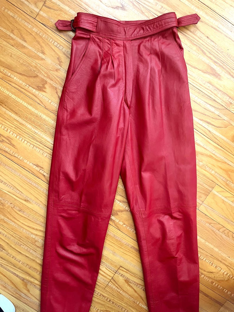 May include: Red leather pants with a high waist and a belt. The pants have pleats at the front and pockets. The leather has a slight sheen. The belt has a buckle closure. The pants are a bold, solid color.