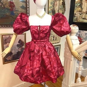 May include: A red floral-patterned dress with a square neckline and puff sleeves is displayed on a mannequin. The dress features a bow at the waist and is paired with a matching head covering. A gold necklace adds a touch of elegance to the ensemble.