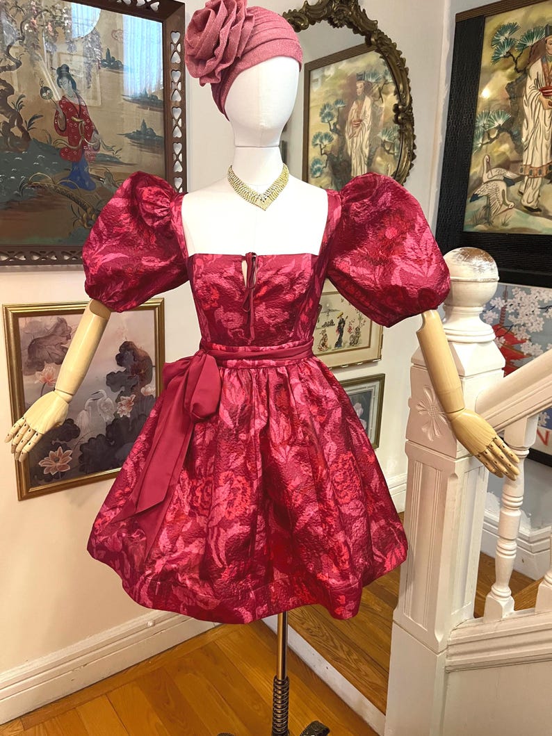 May include: A red floral-patterned dress with a square neckline and puff sleeves is displayed on a mannequin. The dress features a bow at the waist and is paired with a matching head covering. A gold necklace adds a touch of elegance to the ensemble.
