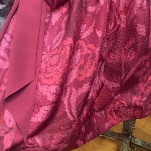 May include: A close-up of a formal dress in shades of magenta and rose. The fabric has a textured, floral pattern. A solid magenta sash is draped over the patterned fabric, creating a layered effect. The dress appears to be made of a luxurious material.