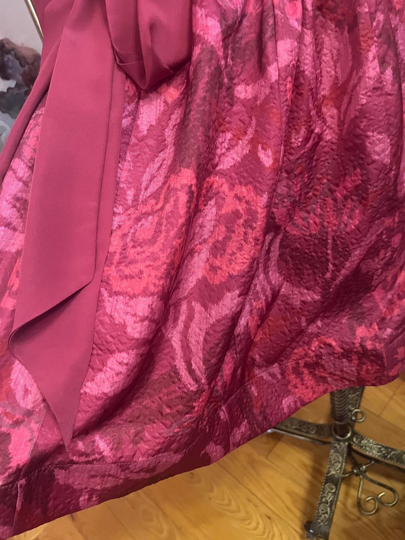 May include: A close-up of a formal dress in shades of magenta and rose. The fabric has a textured, floral pattern. A solid magenta sash is draped over the patterned fabric, creating a layered effect. The dress appears to be made of a luxurious material.