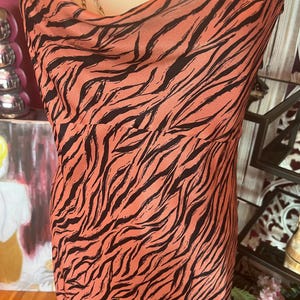 Puede incluir: Vestido sin mangas con cuello de cisne, con fondo coral y rayas de cebra negras. El vestido es de material sedoso y cae suelto. Un collar de cadena dorada es visible en el escote.