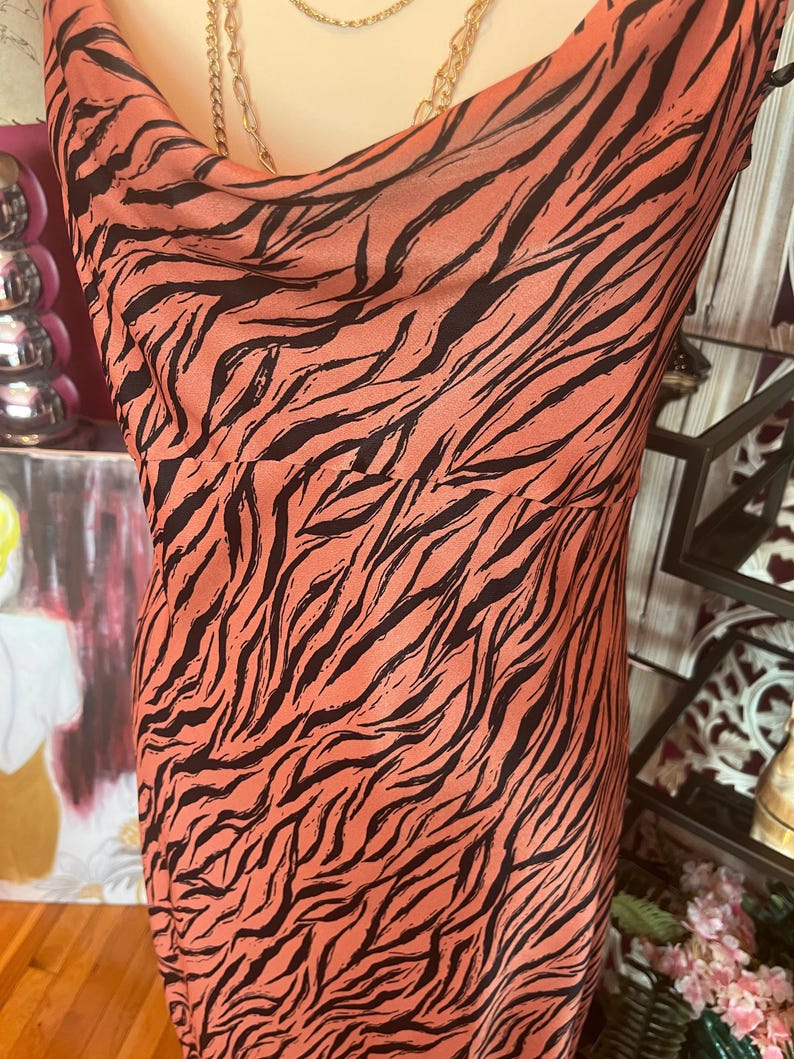 Puede incluir: Vestido sin mangas con cuello de cisne, con fondo coral y rayas de cebra negras. El vestido es de material sedoso y cae suelto. Un collar de cadena dorada es visible en el escote.