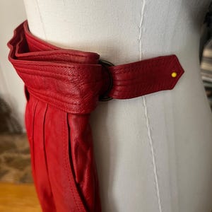 May include: A vibrant red leather belt with a unique design. The belt features a large, folded section and a strap that fastens with a dark metal ring. A yellow pin secures the end of the strap. The belt is displayed on a mannequin.
