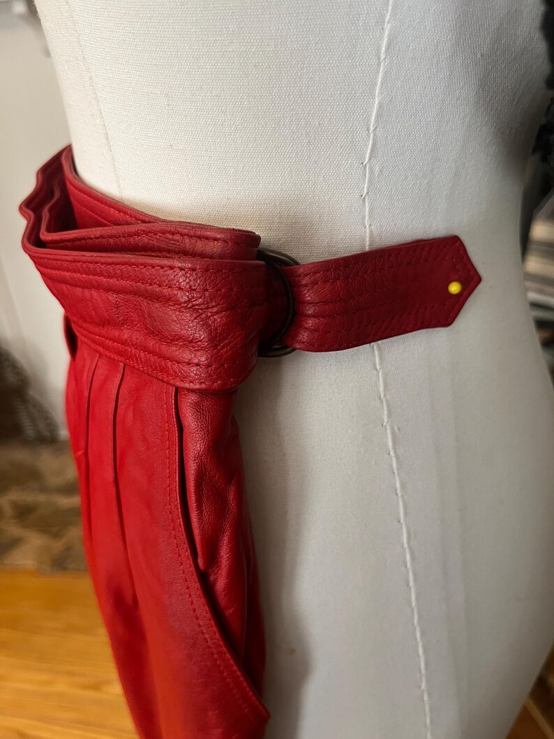 May include: A vibrant red leather belt with a unique design. The belt features a large, folded section and a strap that fastens with a dark metal ring. A yellow pin secures the end of the strap. The belt is displayed on a mannequin.