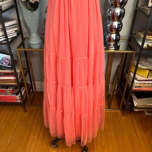 Puede incluir: Un vestido largo color coral con falda escalonada. El vestido presenta un cintur&oacute;n tejido color crema con hebilla marr&oacute;n. El vestido es de tejido ligero y tiene una silueta fluida.