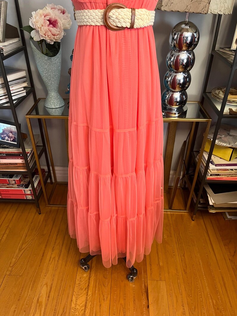 Puede incluir: Un vestido largo color coral con falda escalonada. El vestido presenta un cintur&oacute;n tejido color crema con hebilla marr&oacute;n. El vestido es de tejido ligero y tiene una silueta fluida.