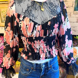 May include: Black floral print blouse with pink flowers and long sleeves. The blouse features a black lace neckline and a cinched waist. The sleeves have a black lace trim at the cuffs. A gold heart pendant necklace is visible.