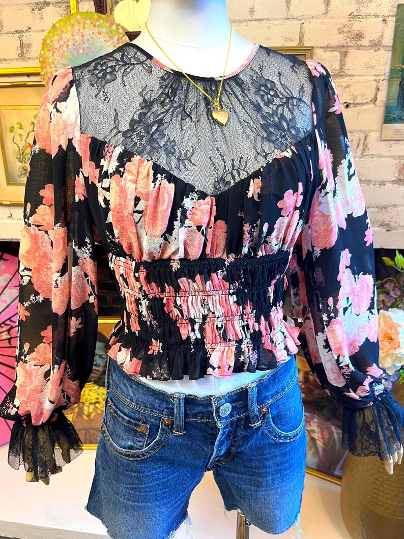 May include: Black floral print blouse with pink flowers and long sleeves. The blouse features a black lace neckline and a cinched waist. The sleeves have a black lace trim at the cuffs. A gold heart pendant necklace is visible.