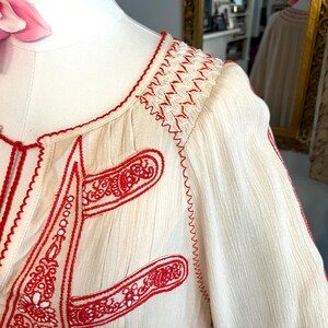 May include: Cream-coloured blouse with intricate red embroidery. The design features detailed stitching along the neckline, sleeves, and front panels. The embroidery includes floral and geometric patterns, creating a bohemian aesthetic. The fabric has a textured appearance.