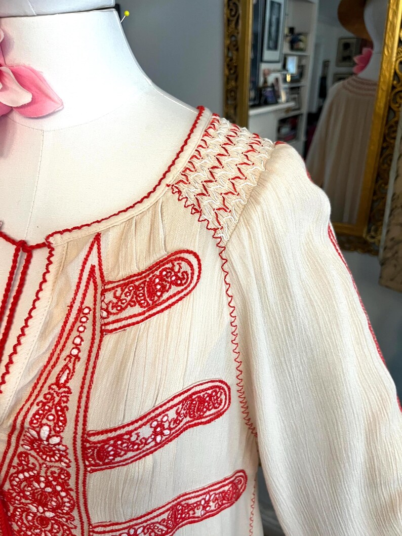 May include: Cream-coloured blouse with intricate red embroidery. The design features detailed stitching along the neckline, sleeves, and front panels. The embroidery includes floral and geometric patterns, creating a bohemian aesthetic. The fabric has a textured appearance.