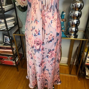 May include: A light pink floral print slip dress with spaghetti straps and a tie-back detail. The dress features a pattern of pink and blue roses. The dress is made of a silky material and has a flowing silhouette.