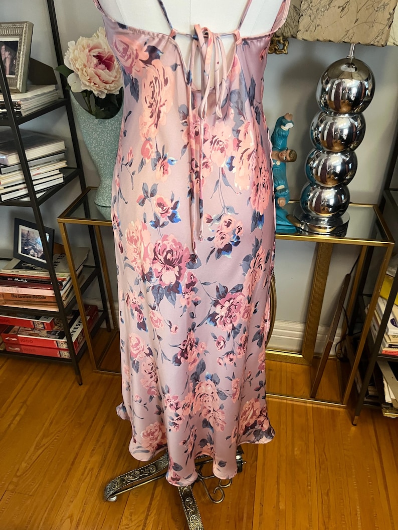May include: A light pink floral print slip dress with spaghetti straps and a tie-back detail. The dress features a pattern of pink and blue roses. The dress is made of a silky material and has a flowing silhouette.