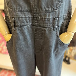 May include: Charcoal gray denim overalls with classic bib pockets, side pockets, and adjustable straps. The overalls have a button closure on the sides and a relaxed fit. A timeless and versatile garment.