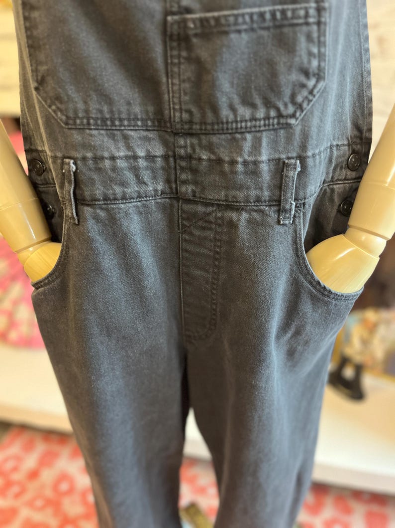 May include: Charcoal gray denim overalls with classic bib pockets, side pockets, and adjustable straps. The overalls have a button closure on the sides and a relaxed fit. A timeless and versatile garment.