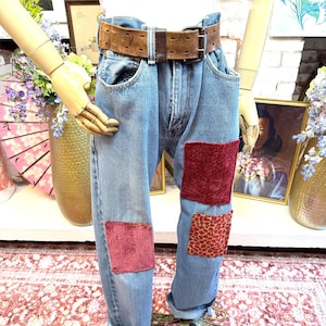 Könnte beinhalten: Hellblaue Jeans aus Denim mit einem braunen Ledergürtel und einer Metallschnalle. Die Hose hat drei rechteckige Flicken auf den Beinen: einen dunkelroten Samtflicken, einen strukturierten rosafarbenen Flicken und einen gemusterten roten Flicken.