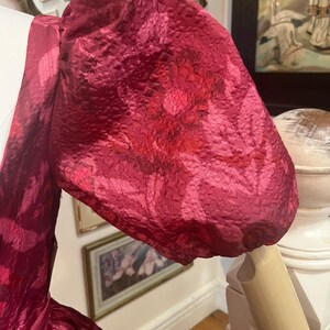 May include: Close-up of a magenta-colored dress sleeve with a floral pattern. The fabric has a textured appearance, and the sleeve is puffed at the shoulder. The dress is displayed on a mannequin, showcasing the design and fabric details.