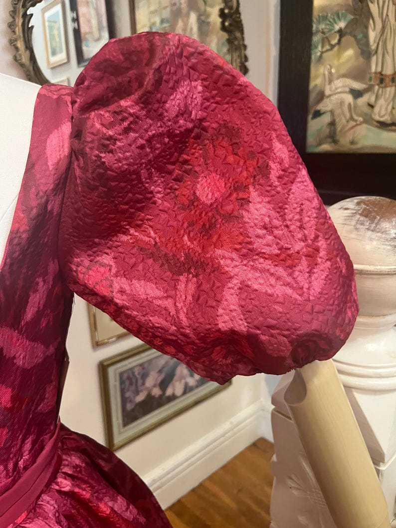 May include: Close-up of a magenta-colored dress sleeve with a floral pattern. The fabric has a textured appearance, and the sleeve is puffed at the shoulder. The dress is displayed on a mannequin, showcasing the design and fabric details.