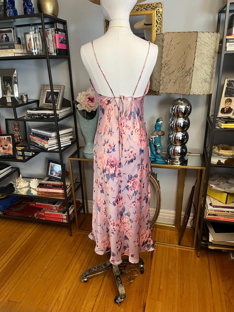 May include: A light pink floral print slip dress on a mannequin. The dress features a delicate floral pattern in shades of pink and blue. The dress has spaghetti straps and a flowing silhouette, perfect for a special occasion.