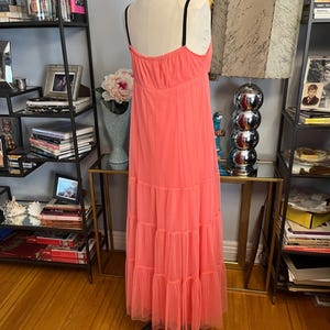 Puede incluir: Un vestido largo color coral con tirantes finos negros se exhibe en un maniqu&iacute;. El vestido presenta una falda escalonada hecha de una tela transparente, creando una silueta fluida. El vestido es una pieza vibrante y llamativa.