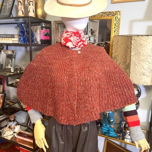 May include: A rust-colored knitted cape with a round neckline. The cape is paired with a cream-colored hat with a brown band. The mannequin is also wearing a red patterned scarf and gray and striped sleeves. The mannequin is wearing brown leather pants.