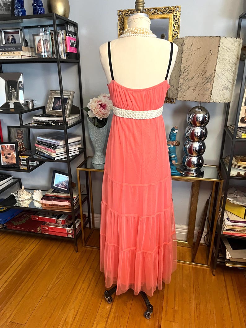 Puede incluir: Vestido largo color coral con cintur&oacute;n de cuerda blanco y tirantes finos negros. El vestido se exhibe en un maniqu&iacute;. El vestido tiene una falda escalonada y est&aacute; hecho de una tela transparente.
