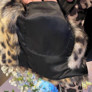 K&ouml;nnte beinhalten: Eine Jacke mit schwarzem Innenfutter aus Stoff und einer Tasche, umrandet von einem Webpelzbesatz mit braun-schwarzem Leopardenmuster. Das Kleidungsst&uuml;ck zeigt ein strukturiertes Tiermuster an den Au&szlig;enkanten und an den &Auml;rmeln.