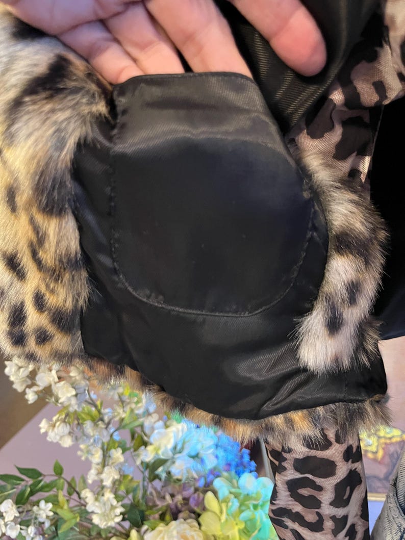 K&ouml;nnte beinhalten: Eine Jacke mit schwarzem Innenfutter aus Stoff und einer Tasche, umrandet von einem Webpelzbesatz mit braun-schwarzem Leopardenmuster. Das Kleidungsst&uuml;ck zeigt ein strukturiertes Tiermuster an den Au&szlig;enkanten und an den &Auml;rmeln.