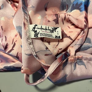 May include: Close-up of a silky, floral-patterned garment in shades of pink, blue, and peach. The fabric is draped, revealing a tag that reads "Intimately Free People". The garment appears to be a top or a dress.