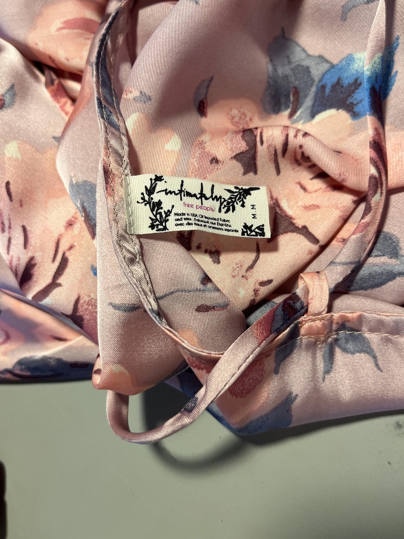May include: Close-up of a silky, floral-patterned garment in shades of pink, blue, and peach. The fabric is draped, revealing a tag that reads "Intimately Free People". The garment appears to be a top or a dress.
