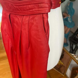 May include: Bright red leather pants with a wide, belted waistband. The pants have a high waist and pleats at the front. The belt has a buckle closure. The pants are displayed on a mannequin.