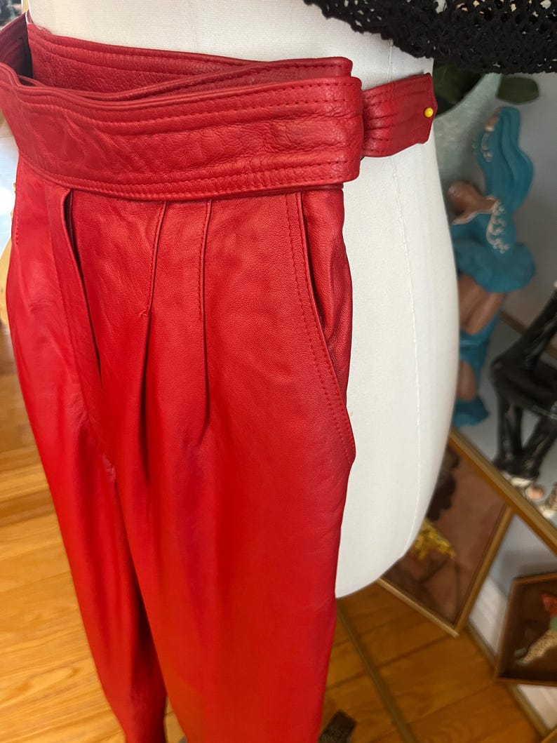 May include: Bright red leather pants with a wide, belted waistband. The pants have a high waist and pleats at the front. The belt has a buckle closure. The pants are displayed on a mannequin.
