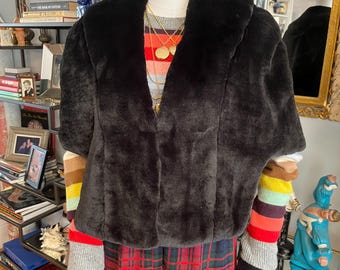 Puede incluir: Una chaqueta negra de piel sintética con cuello ancho se exhibe en un maniquí. La chaqueta se combina con un suéter a rayas, pantalones a cuadros y guantes negros. Los collares dorados añaden un toque de elegancia al conjunto. El estilo general es una mezcla de texturas y patrones.