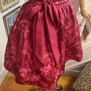 May include: A burgundy-colored dress with a patterned design and a matching sash tied in a bow. The dress is displayed on a vintage dress form. The fabric has a textured appearance.
