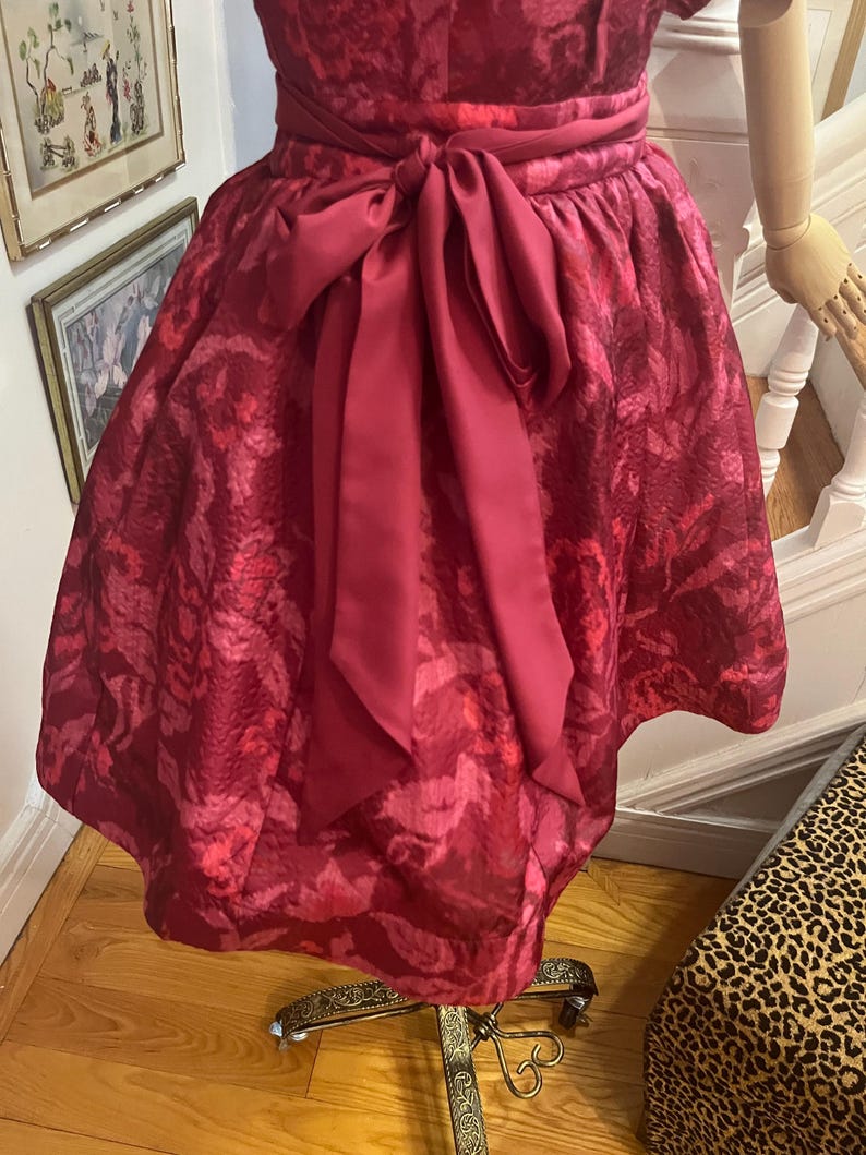 May include: A burgundy-colored dress with a patterned design and a matching sash tied in a bow. The dress is displayed on a vintage dress form. The fabric has a textured appearance.