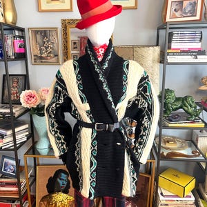 Puede incluir: Un maniquí con un sombrero fedora rojo con una banda marrón, una rebeca estampada en negro y crema, un cinturón negro y pantalones de cuadros rojos. La rebeca tiene detalles en verde. Gafas de sol metidas en el cinturón. El fondo incluye obras de arte enmarcadas y estanterías.