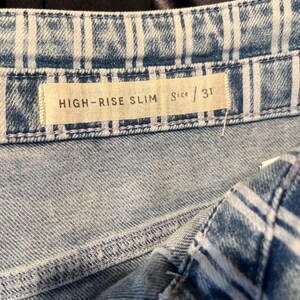 Pode incluir: Close-up de jeans de ganga azul com corte slim de cintura alta. Os jeans t&ecirc;m uma lavagem clara com riscas verticais brancas. A etiqueta de tamanho diz "HIGH-RISE SLIM" e "Size / 31".