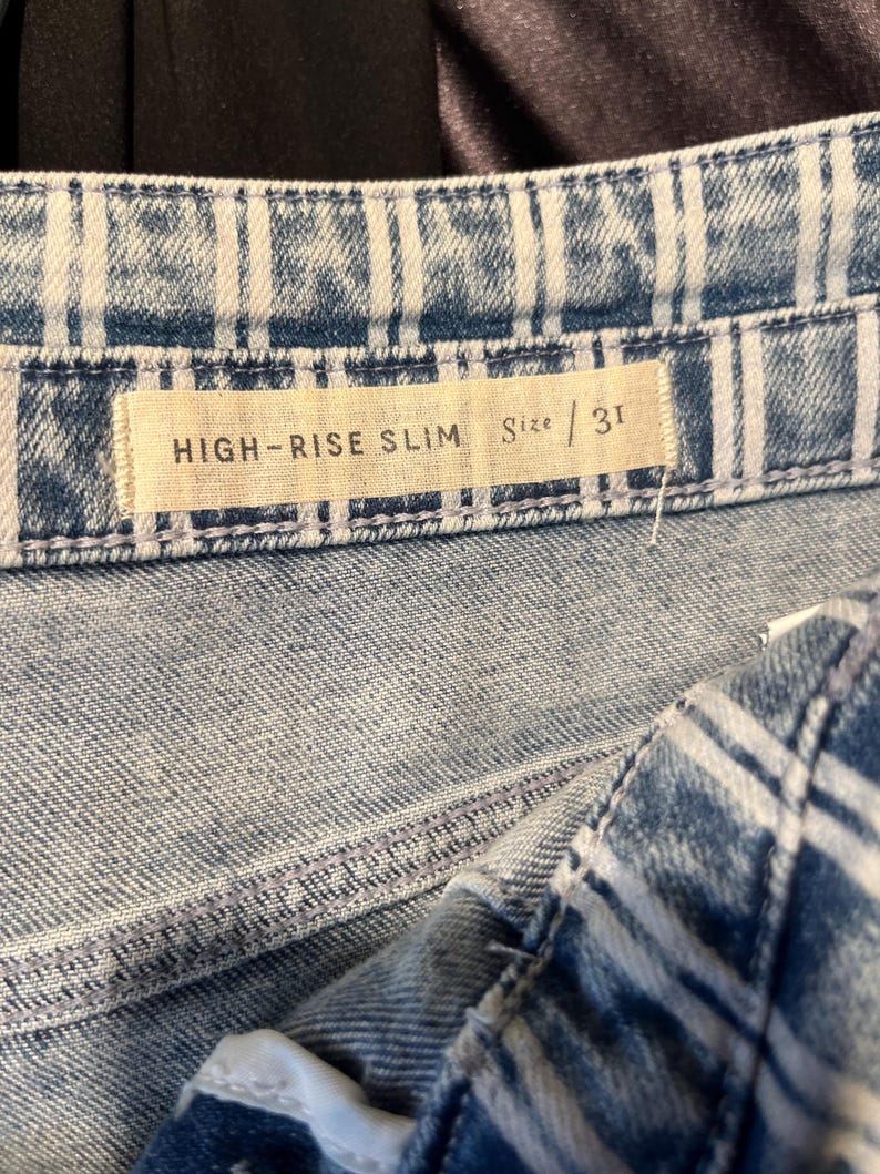 Pode incluir: Close-up de jeans de ganga azul com corte slim de cintura alta. Os jeans t&ecirc;m uma lavagem clara com riscas verticais brancas. A etiqueta de tamanho diz "HIGH-RISE SLIM" e "Size / 31".