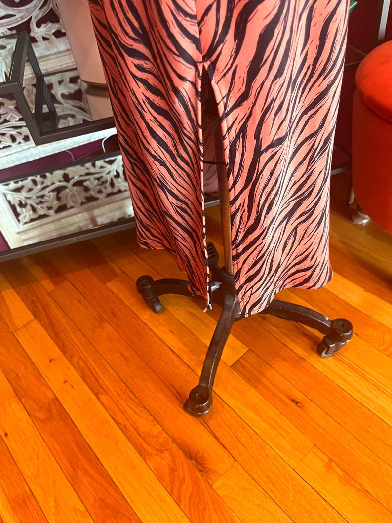 Puede incluir: Un vestido largo con un vibrante fondo coral y audaces rayas de cebra negras. El vestido se exhibe en un maniqu&iacute; de metal negro con ruedas, mostrando su dise&ntilde;o y silueta. El vestido tiene una abertura frontal.