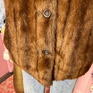 May include: A close-up of a brown fur vest with two dark brown buttons. The vest has a soft, textured appearance and a classic design. The fur appears to be in good condition.