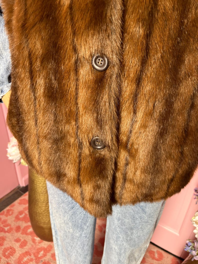 May include: A close-up of a brown fur vest with two dark brown buttons. The vest has a soft, textured appearance and a classic design. The fur appears to be in good condition.