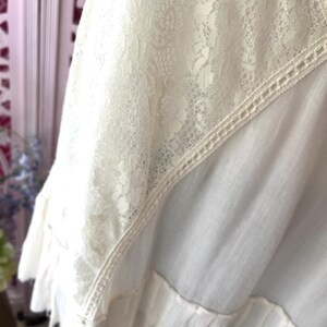 Op de afbeelding: Close-up van een cr&egrave;mekleurig kledingstuk met een delicaat kanten paneel en een gefranjerde zoom. Het kantdetail is ingewikkeld bewerkt en de stof lijkt zacht en vloeiend. Het kledingstuk heeft een vintage of bohemien esthetiek.