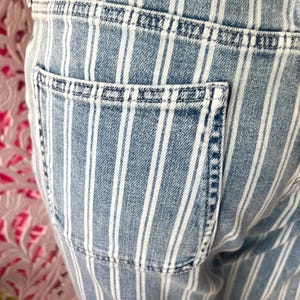 Pode incluir: Close-up de jeans de ganga azul claro com riscas brancas verticais. Os jeans apresentam um bolso traseiro e detalhes de costura vis&iacute;veis. O tecido tem um aspeto desbotado e gasto.